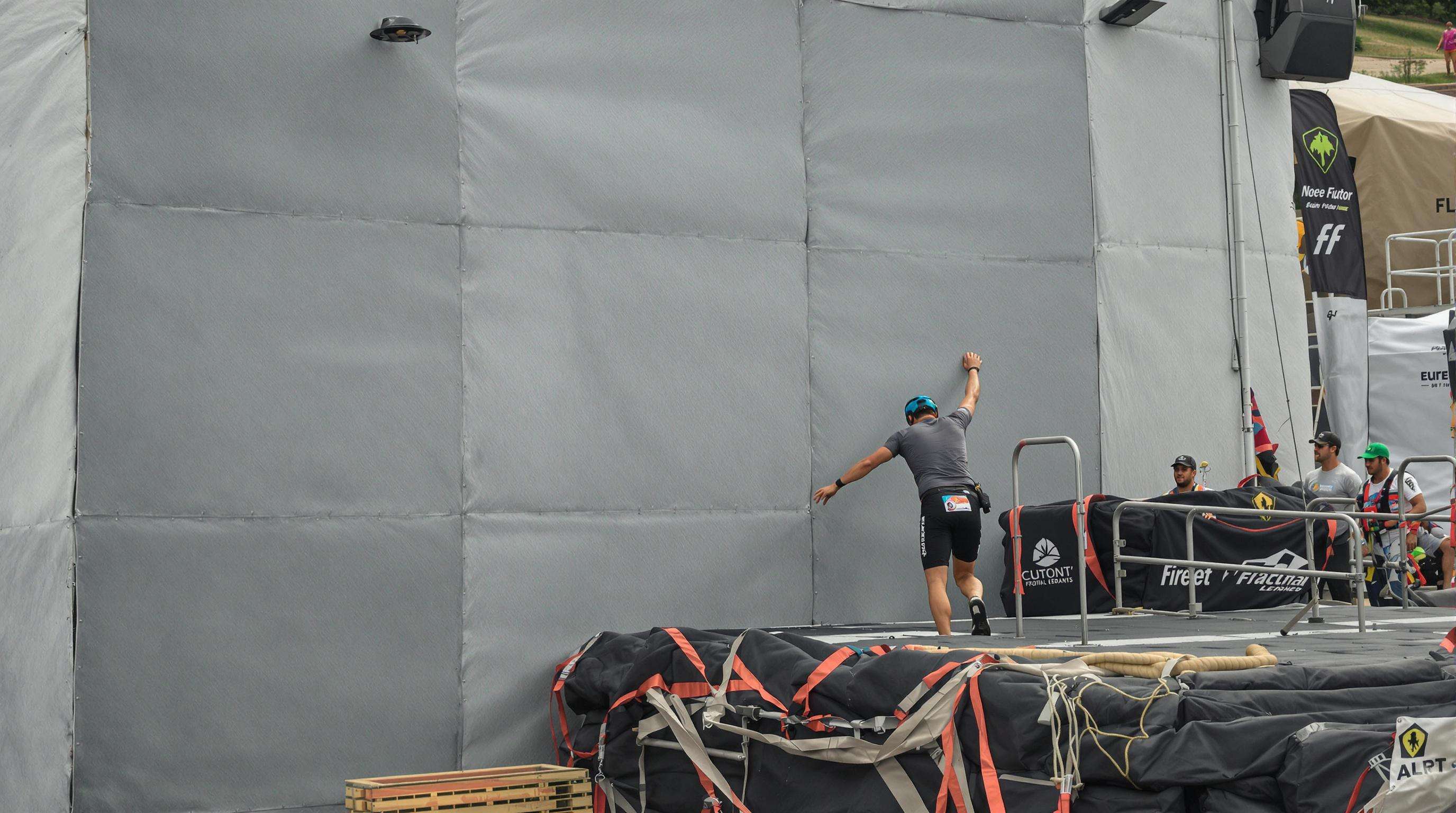 Participant lands safely on a foam-padded surface next to a tall obstacle while staff monitor for safety