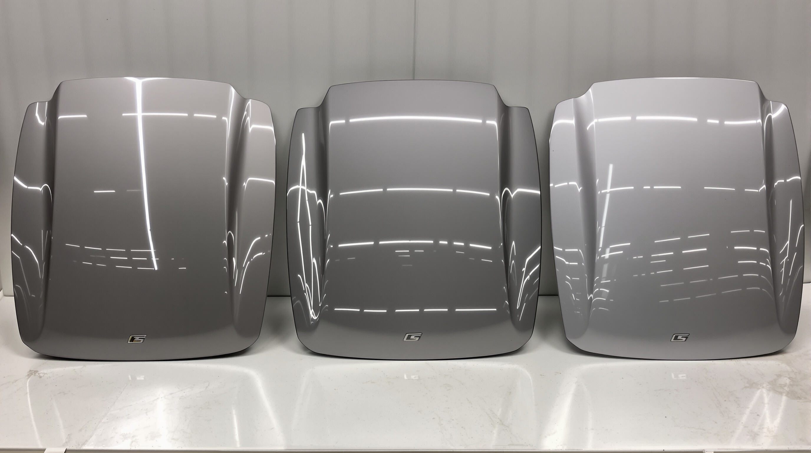 Three car trunk lids made of steel, aluminum, and plastic composite displayed side by side in a muted workshop setting
