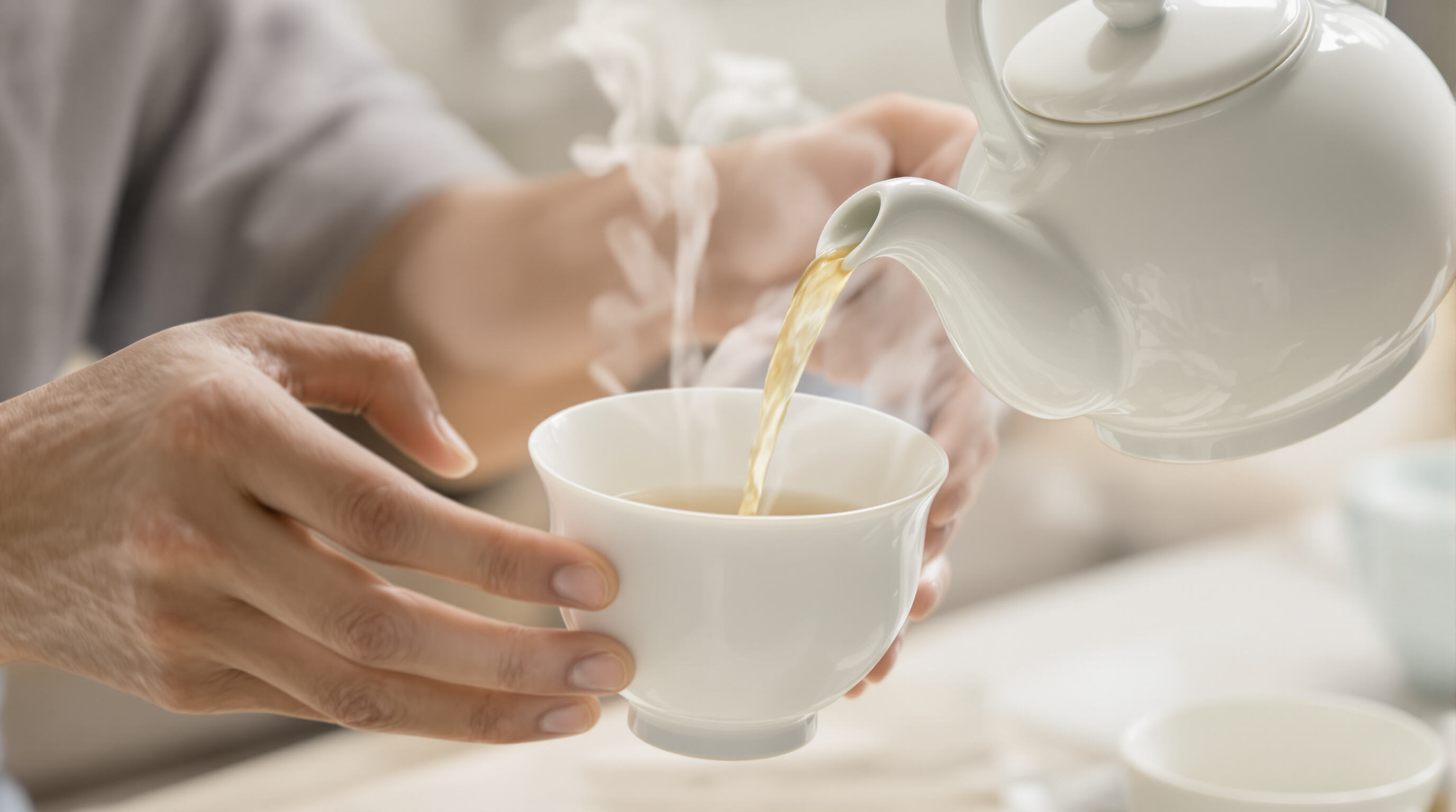 Close-up of hands pouring tea from an ergonomic Chinese porcelain set with curved cups and steam