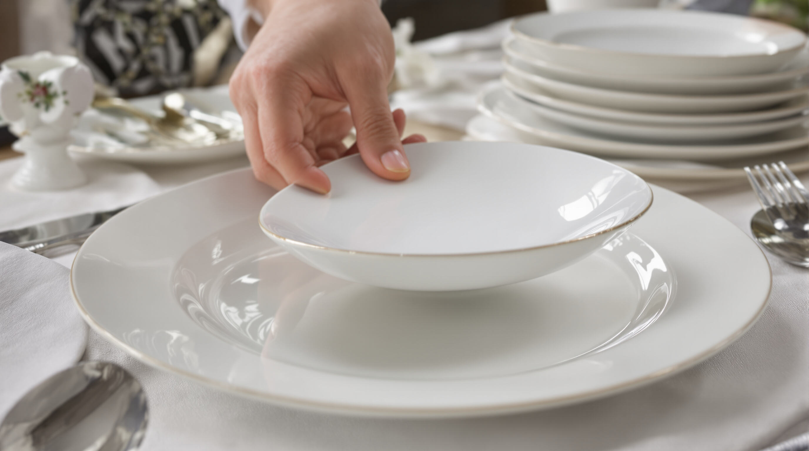 Hand lifting a thin bone china plate, showing glazed rim and lightweight design amid a soft-toned table setting