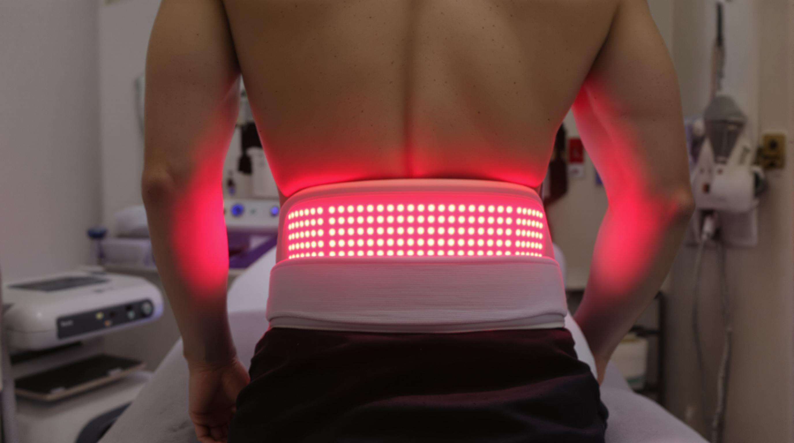 Photorealistic image of a red light therapy belt emitting gentle red and infrared light on a person's lower back in a clinical environment