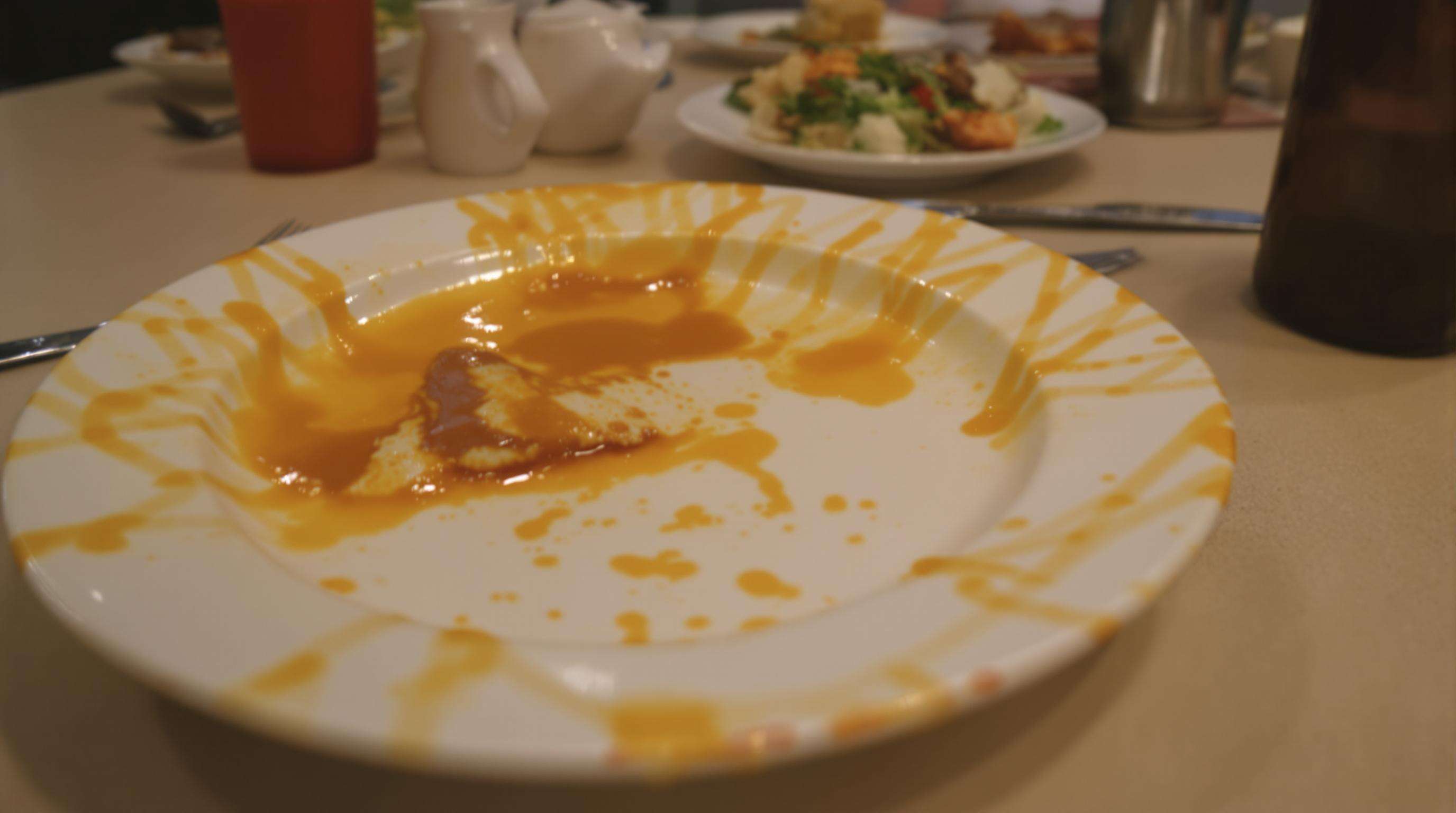 Shallow ceramic dinner plate with food and sauce spilling over the edge onto a table.