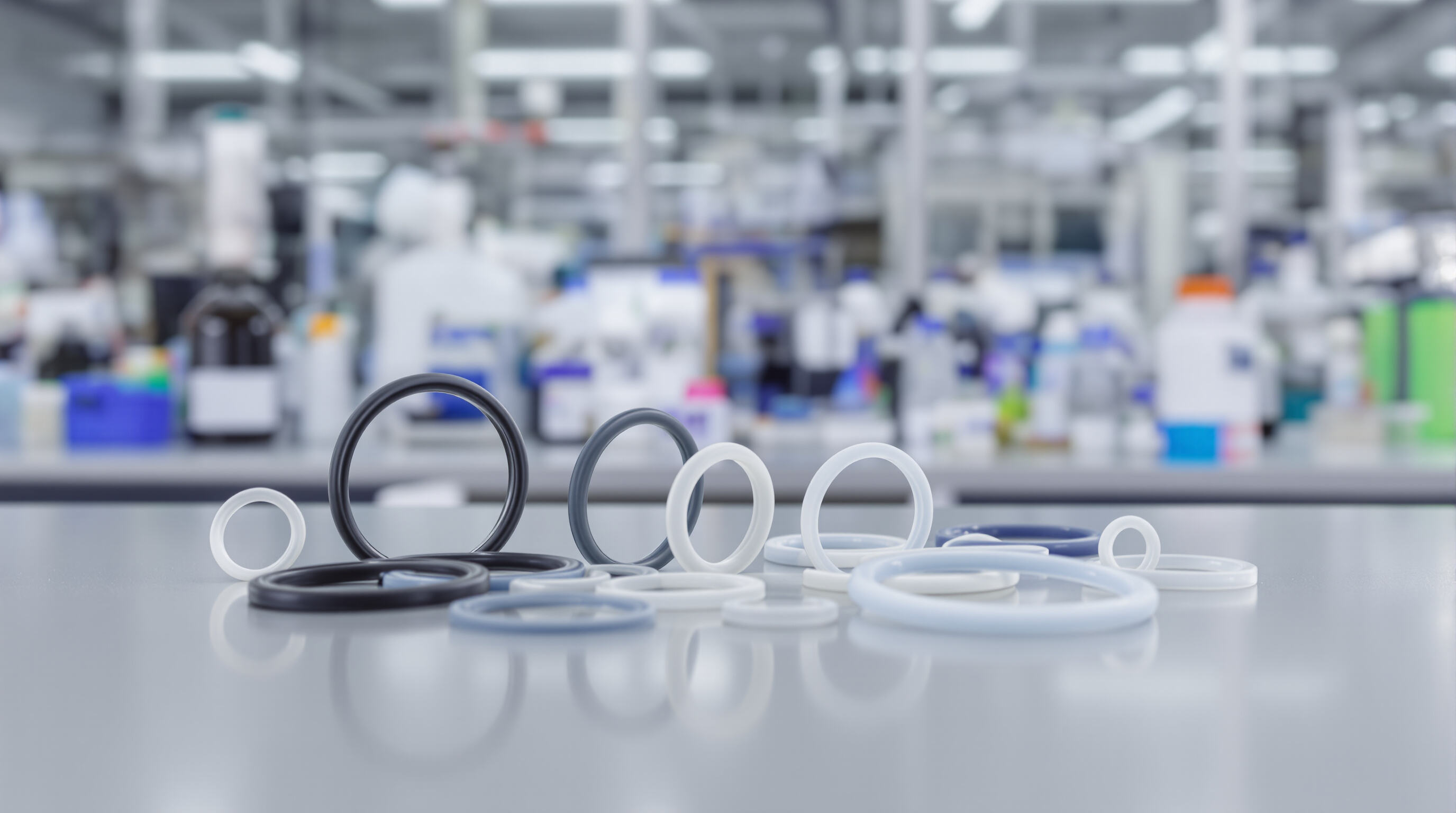 Close-up of various o-rings made from different materials on a lab bench with industrial equipment blurred in the background
