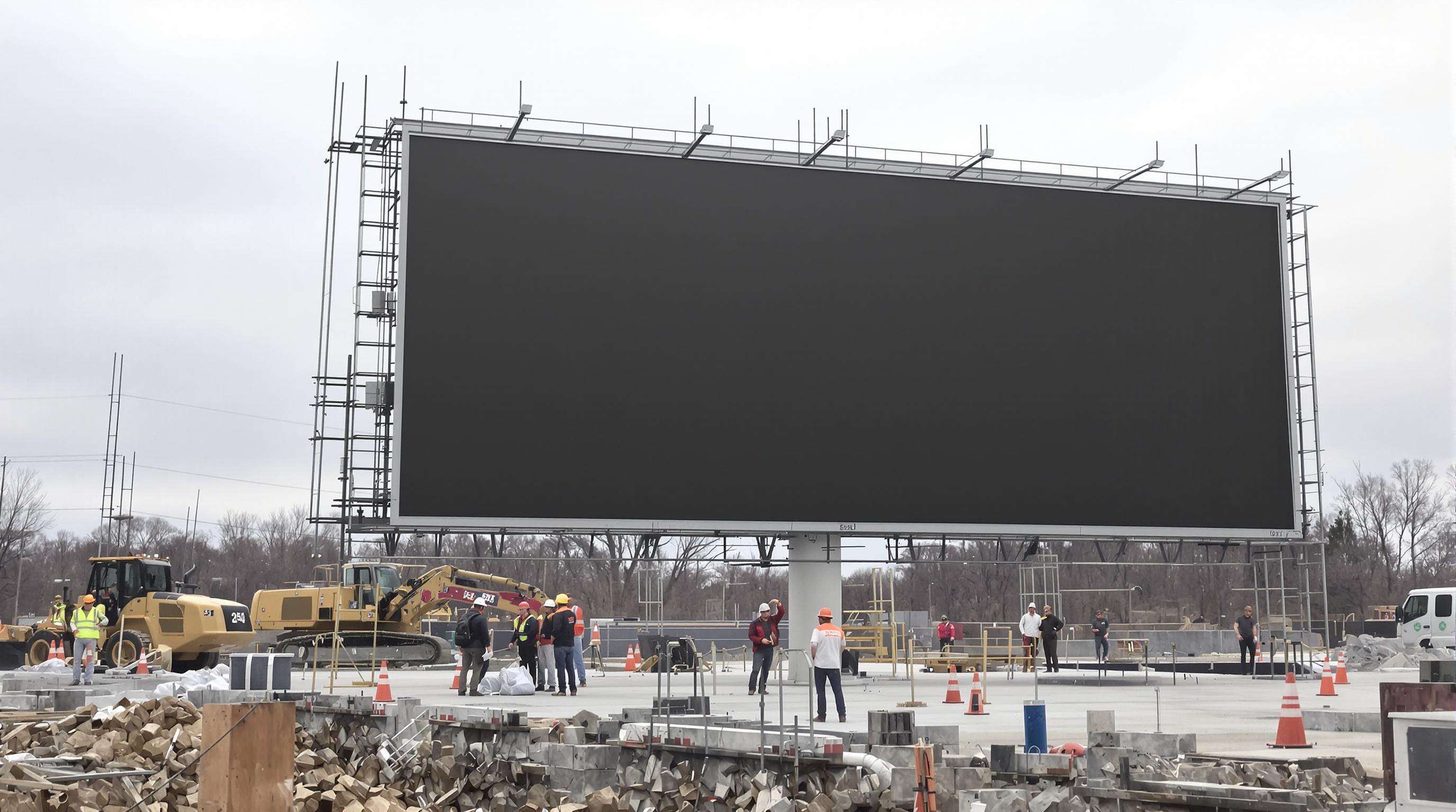 Photo of workers and machines installing a large digital billboard, with visible foundation and terrain challenges