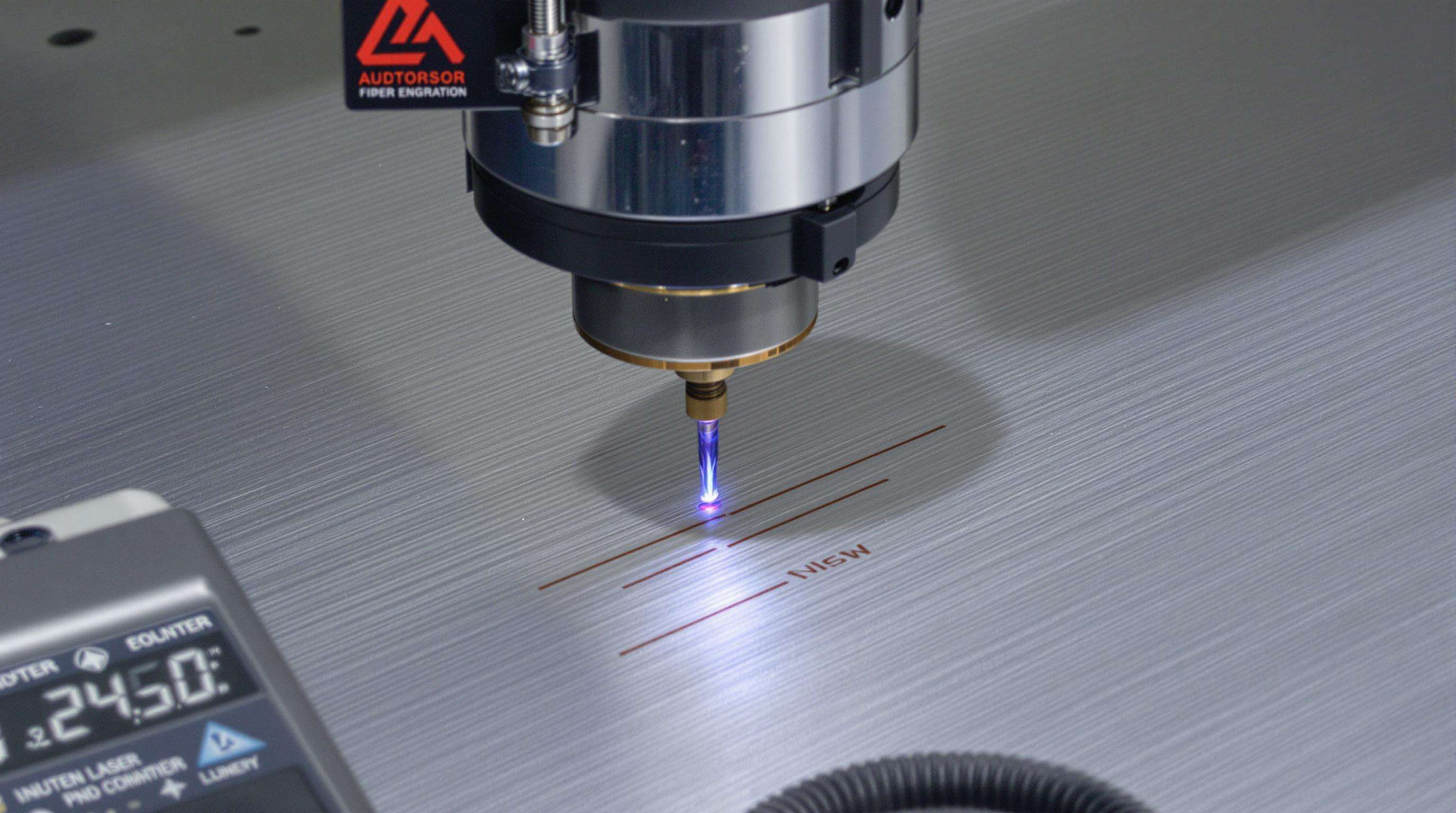 Close-up of a fiber laser engraver marking an aluminum surface, with dirty lens and multimeter nearby.
