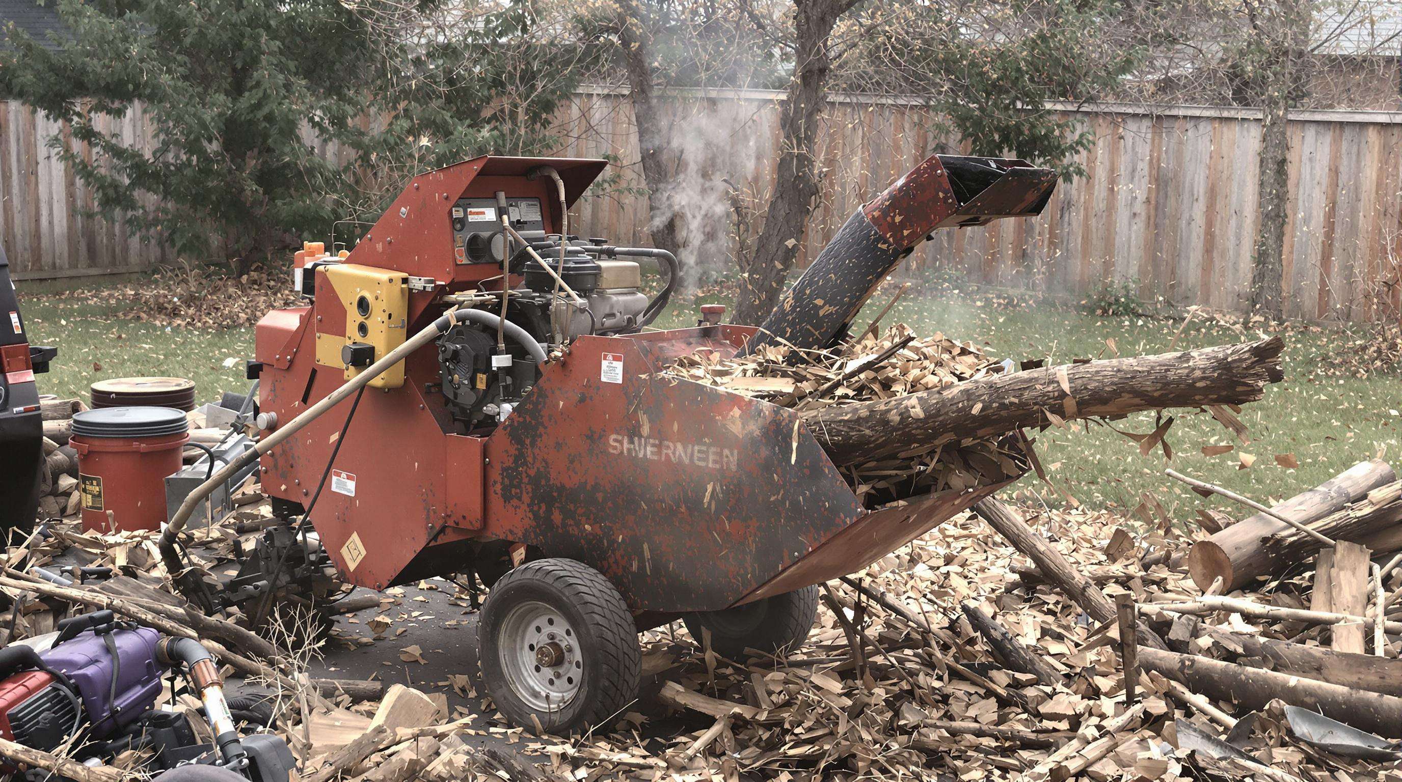 An older chipper shredder in a backyard having difficulty processing a branch, with an operator nearby and wood debris piling up