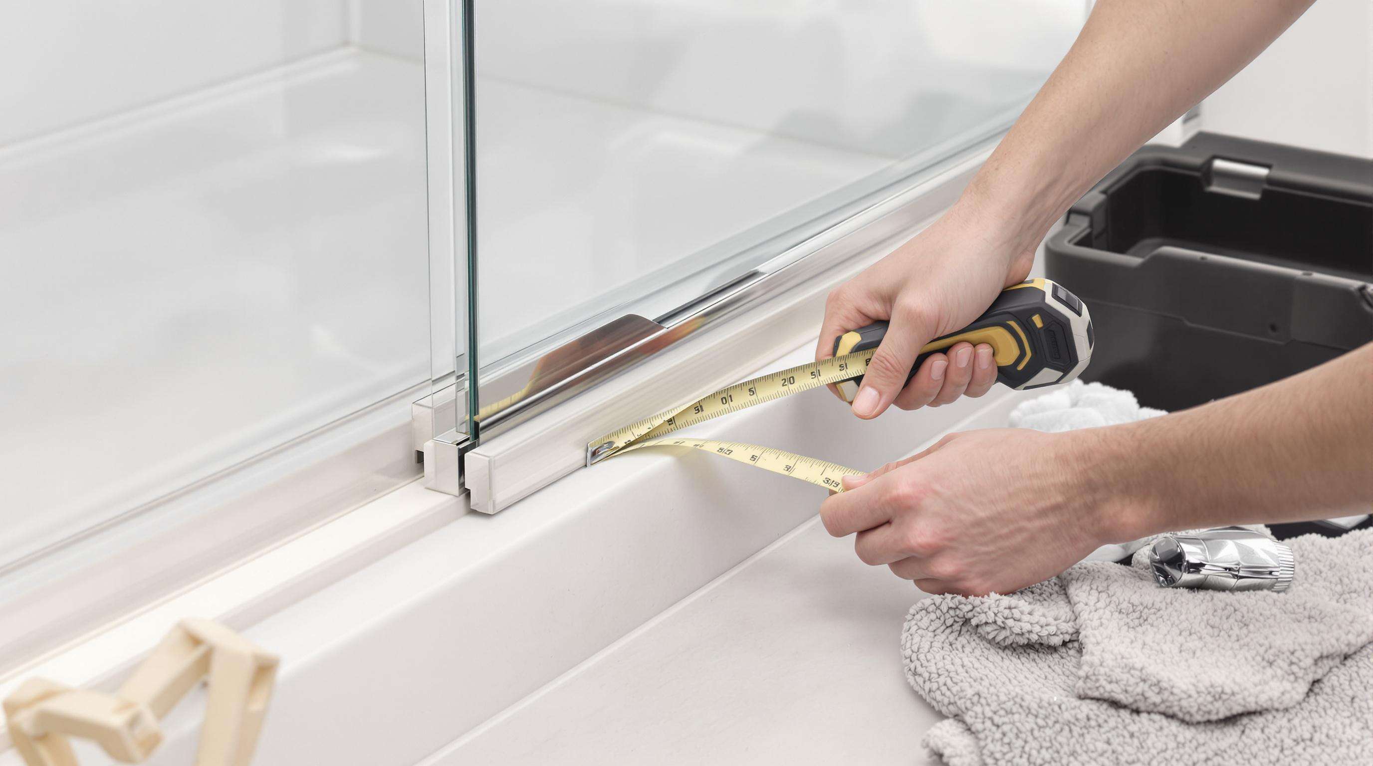 Hands measuring and cutting a shower door seal beside a glass panel with tools nearby