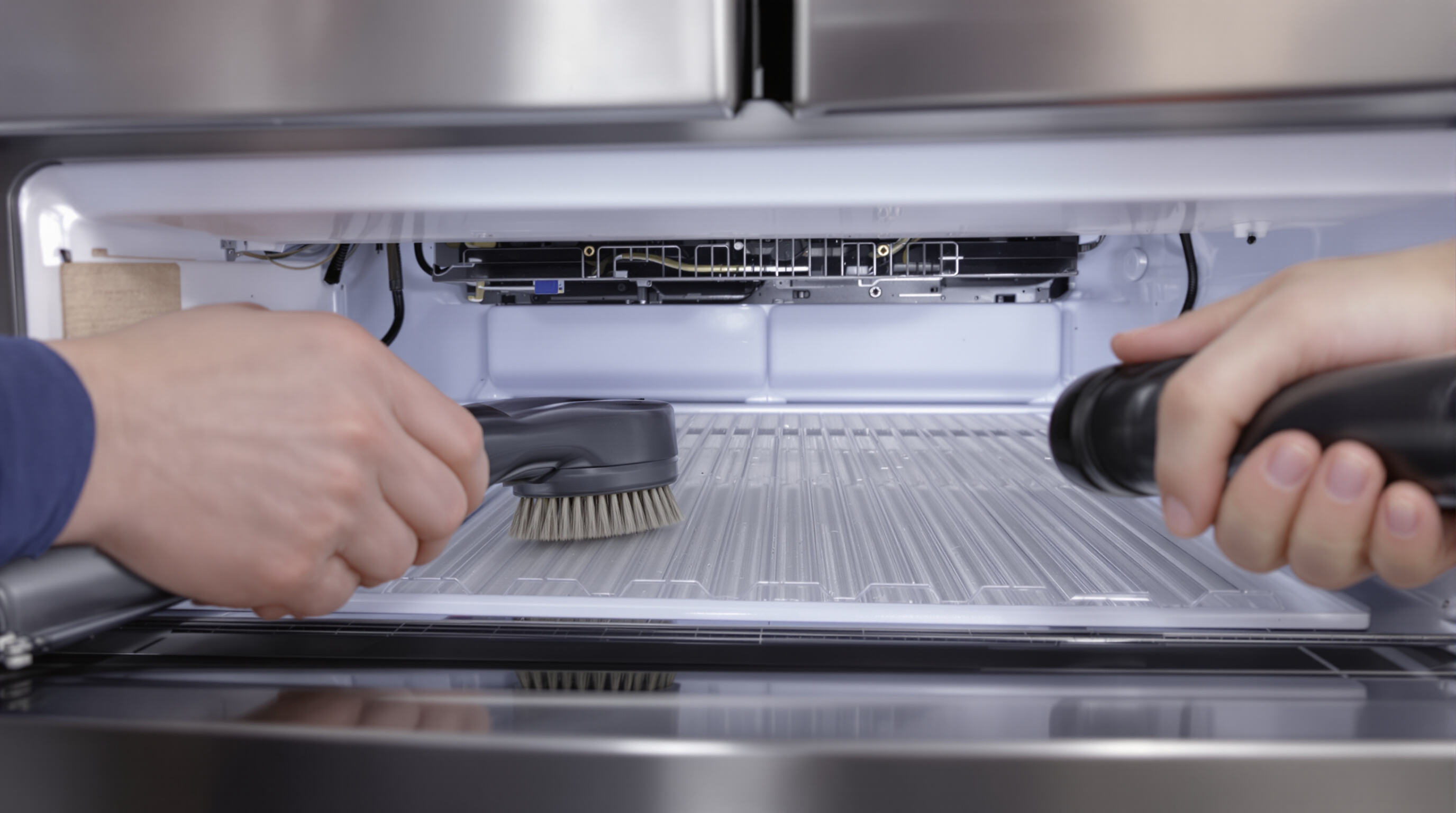 Technician cleaning dust from refrigerator condenser coils with brush and vacuum