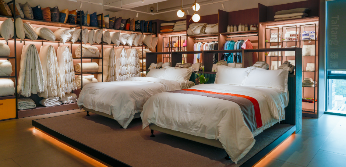 A photograph of two modern hotel beds with clean-line Taitang brand bedding and LED under-lighting, with a background wall of meticulously arranged, luxurious bedding collections, pillows, and linens in a professional showroom display.