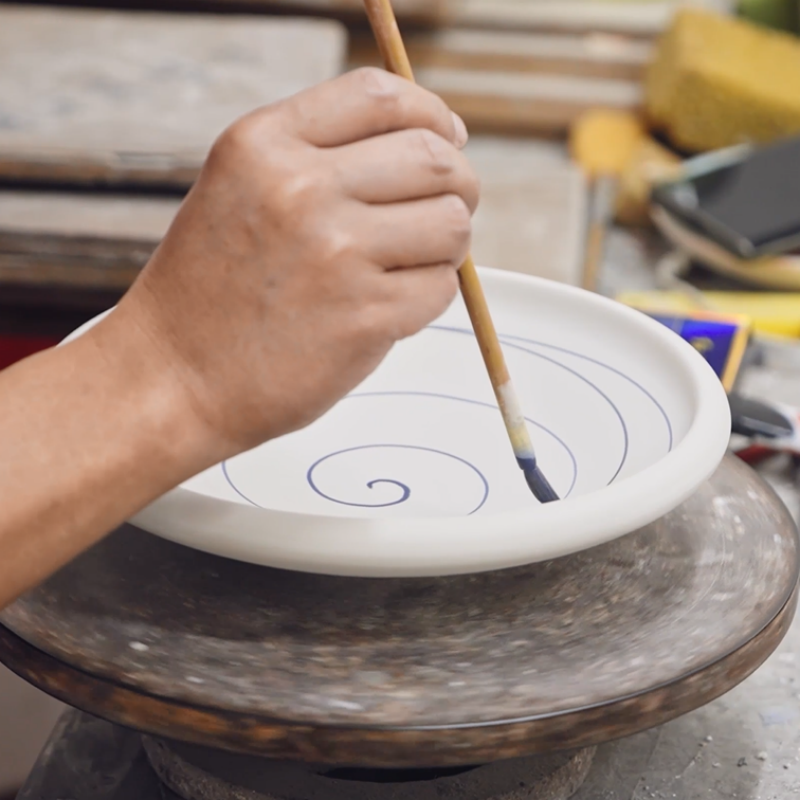 Close-up of a skilled factory artisan hand-painting custom patterns onto a white ceramic plate on a decorating wheel..png