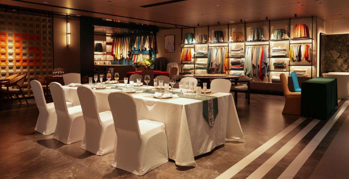 A professionally arranged banquet setup within a Taitang showroom, featuring a long dining table with a crisp white tablecloth, matching chair covers, and sparkling glassware, positioned in front of a comprehensive and brightly lit display of colorful dining textiles and hotel furniture options for events.
