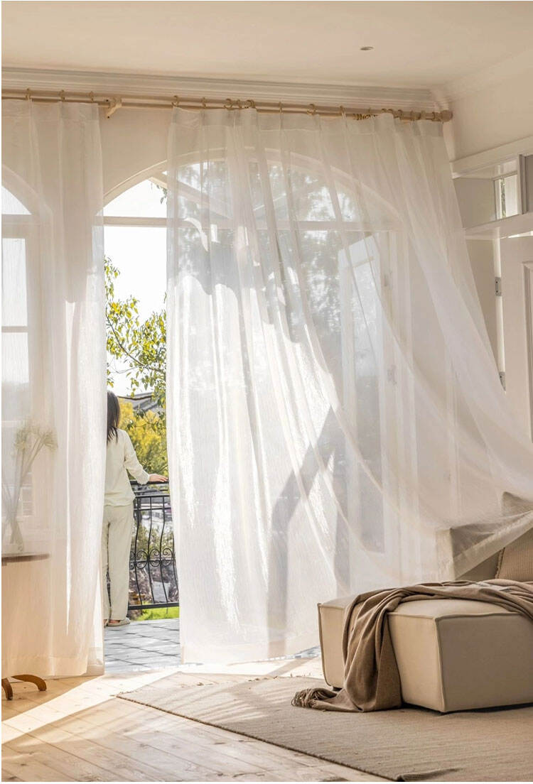 Sunlit living room with flowing white glass sheer curtains by an arched balcony door, diffusing light while keeping privacy.